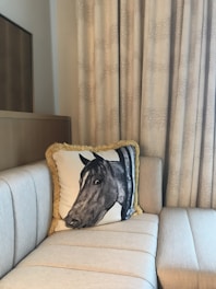 A beige cushioned sofa in the corner of a room with a decorative pillow featuring a horse's head. Behind the sofa are light beige curtains with a subtle patterned design. A wooden cabinet with a reflective surface is partially visible in the background.