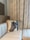 A beige cushioned sofa in the corner of a room with a decorative pillow featuring a horse's head. Behind the sofa are light beige curtains with a subtle patterned design. A wooden cabinet with a reflective surface is partially visible in the background.