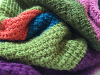 A close-up view of a knitted fabric featuring a variety of textured patterns in different colors, including green, orange, blue, pink, and purple. The fabric appears soft and cozy, highlighting the intricate weave and craftsmanship.