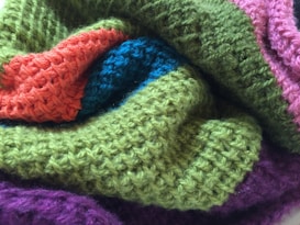 A close-up view of a knitted fabric featuring a variety of textured patterns in different colors, including green, orange, blue, pink, and purple. The fabric appears soft and cozy, highlighting the intricate weave and craftsmanship.