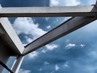 A white concrete structure is set against a bright blue sky scattered with fluffy clouds. The angular architecture contrasts sharply with the softness of the clouds, creating a striking interplay between natural and man-made elements.