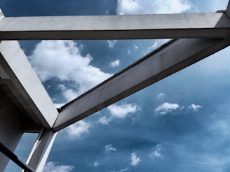 A white concrete structure is set against a bright blue sky scattered with fluffy clouds. The angular architecture contrasts sharply with the softness of the clouds, creating a striking interplay between natural and man-made elements.