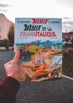 A hand holds an Asterix comic book titled 'Asterix et la Transitalique.' The cover features colorful illustrations of Asterix characters in a dynamic chariot race scene with vibrant and exaggerated expressions.