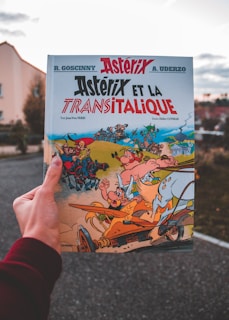 A hand holds an Asterix comic book titled 'Asterix et la Transitalique.' The cover features colorful illustrations of Asterix characters in a dynamic chariot race scene with vibrant and exaggerated expressions.