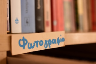 A bookshelf is in focus with several books in various colors positioned upright. Nearby, a label with blue handwriting in a non-Latin script is attached to the shelf edge.
