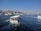 A large passenger boat navigates through a wide waterway with a cityscape in the background. The skyline is adorned with historic architecture, including several domes and minarets. The water is a deep blue and multiple boats can be seen on the water. There is a bridge with a modern design spanning the water in the distance.