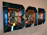 Intricate geometric mirrors with a three-dimensional effect are mounted on a wall, reflecting parts of an interior space. The mirrors create a kaleidoscopic pattern with varying reflections and angles.