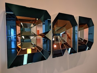 Intricate geometric mirrors with a three-dimensional effect are mounted on a wall, reflecting parts of an interior space. The mirrors create a kaleidoscopic pattern with varying reflections and angles.