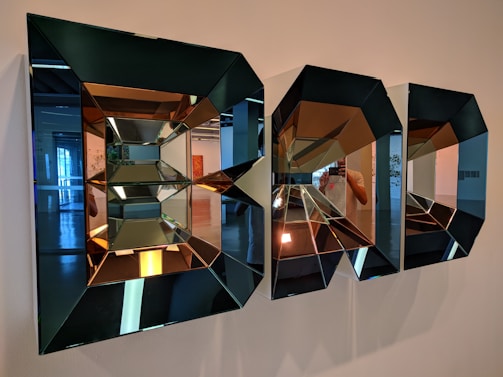 Intricate geometric mirrors with a three-dimensional effect are mounted on a wall, reflecting parts of an interior space. The mirrors create a kaleidoscopic pattern with varying reflections and angles.