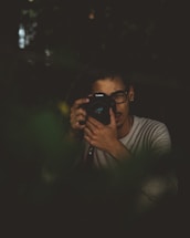 A person with dark-rimmed glasses is holding a camera up to their face, appearing to take a photograph or review images. The setting is dimly lit, with a moody and subdued atmosphere, possibly surrounded by foliage or in a natural environment.