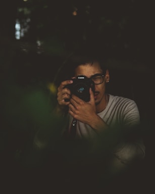 A person with dark-rimmed glasses is holding a camera up to their face, appearing to take a photograph or review images. The setting is dimly lit, with a moody and subdued atmosphere, possibly surrounded by foliage or in a natural environment.