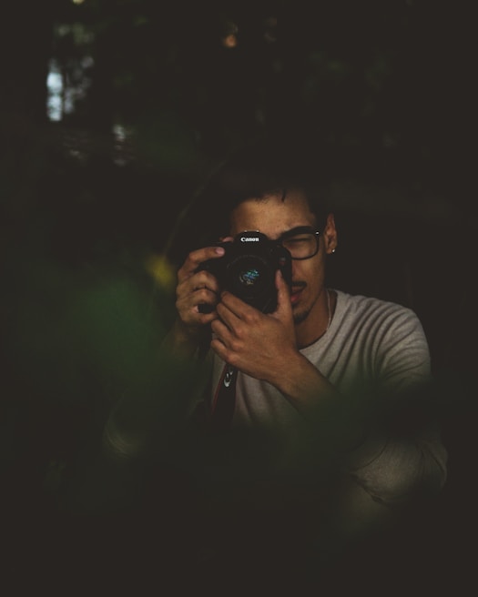 A person with dark-rimmed glasses is holding a camera up to their face, appearing to take a photograph or review images. The setting is dimly lit, with a moody and subdued atmosphere, possibly surrounded by foliage or in a natural environment.