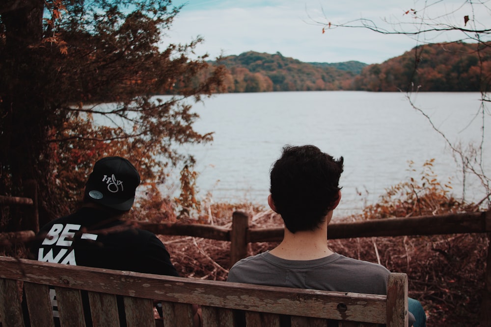 two men sitting on wooden bench looking at body of water