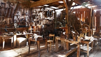 A rustic, old-fashioned workshop filled with vintage tools and wooden furniture. Sunlight filters through the wooden slats, illuminating various hand tools and equipment hung on the walls and scattered on workbenches. The scene suggests a historical or artisanal setting.