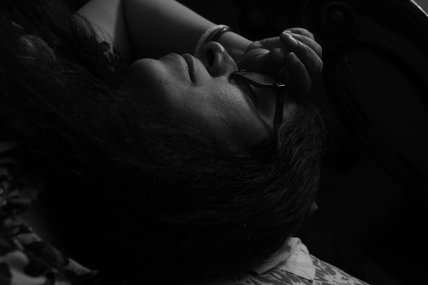 A person is lying down, facing upwards in a dimly lit environment. Their hand is resting on their forehead, and they are wearing glasses. The focus is on the calmness and introspective moment.