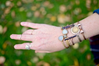 A hand adorned with various bracelets and rings is displayed against a blurred green and brown background. The hand has multiple bracelets featuring stones, metallic elements, and a vintage coin design. The fingers have several rings with unique designs.