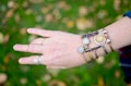 A hand adorned with various bracelets and rings is displayed against a blurred green and brown background. The hand has multiple bracelets featuring stones, metallic elements, and a vintage coin design. The fingers have several rings with unique designs.