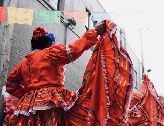 A person wearing a bright orange traditional dress adorned with colorful stripes and lace trim holds up the dress in a celebratory manner. Flowers are tucked into their hair, complementing the vibrant outfit. Above are colorful papel picado banners strung across a narrow alleyway lined with brick walls.