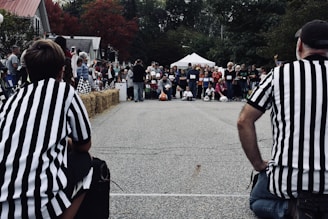 A crowd of people is gathered around an outdoor race track. Two referees wearing black and white striped shirts are positioned at either side of the image, facing the crowd. There are hay bales lined up at the edge of the race track, and some people are sitting on them. The audience appears to be observing an event, possibly a race, with some participants in the background prepared to begin. Trees with fall-colored leaves and a white tent are visible in the background.