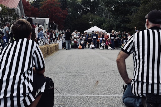 A crowd of people is gathered around an outdoor race track. Two referees wearing black and white striped shirts are positioned at either side of the image, facing the crowd. There are hay bales lined up at the edge of the race track, and some people are sitting on them. The audience appears to be observing an event, possibly a race, with some participants in the background prepared to begin. Trees with fall-colored leaves and a white tent are visible in the background.