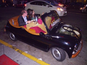 A small black car transformed into an unconventional dining setting, with two people seated inside enjoying a meal. The car's interior is fitted like a cozy booth with cushions and a wooden table, complete with tablecloths, wine glasses, and bottled water. The car hood features an airbrushed image of the Mona Lisa, and the vehicle is parked on a city street with traffic nearby.