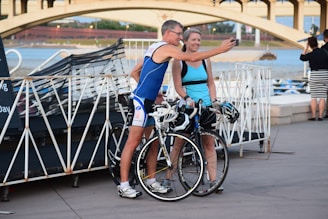 A man and woman stand next to each other with their bicycles, both wearing athletic gear. They appear to be taking a selfie with the man holding the phone. In the background, there is a bridge and a body of water, suggesting they are near a waterfront area.