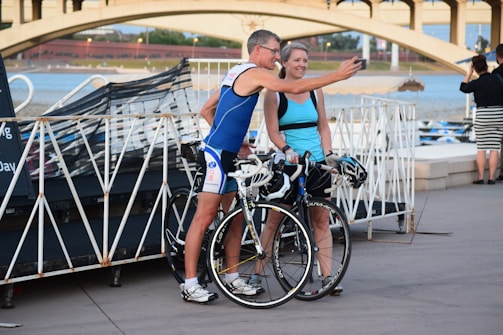 A man and woman stand next to each other with their bicycles, both wearing athletic gear. They appear to be taking a selfie with the man holding the phone. In the background, there is a bridge and a body of water, suggesting they are near a waterfront area.
