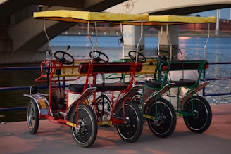 Three colorful four-wheeled pedal bikes with yellow canopies are parked side by side on a paved area by a body of water, with a bridge in the background. The bikes feature steering wheels, black seats, and bars, painted in red, blue, and green.