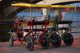 Three colorful four-wheeled pedal bikes with yellow canopies are parked side by side on a paved area by a body of water, with a bridge in the background. The bikes feature steering wheels, black seats, and bars, painted in red, blue, and green.