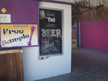 A chalkboard sign with humorous writing is displayed outside a colorful storefront. The sign has playful text about wishing someone was beer, referring to it as an adult daycare center. There is also a sign offering free samples of New Orleans frozen daiquiris. The environment appears casual and light-hearted.