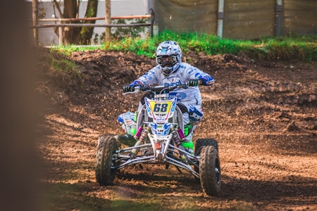 A person is riding a quad bike on a dirt track, kicking up dust. They are wearing a full protective racing suit and helmet, displaying various sponsor logos. The environment appears to be outdoors with some grassy sections and a fence in the background.