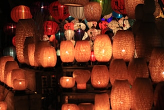 A collection of decorative lanterns in various colors and shapes fills the space, providing a warm and inviting glow. The foreground features numerous woven rattan lanterns emitting a soft orange light, while colorful paper lanterns of red, green, blue, and white are visible in the background.