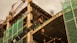 A construction site featuring a partially built structure with exposed concrete and scaffolding. Green mesh netting covers sections of the building for safety. A large crane is visible in the background against an overcast sky, and various building materials and equipment are scattered around.
