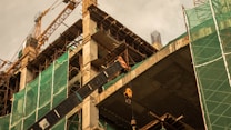 A construction site featuring a partially built structure with exposed concrete and scaffolding. Green mesh netting covers sections of the building for safety. A large crane is visible in the background against an overcast sky, and various building materials and equipment are scattered around.