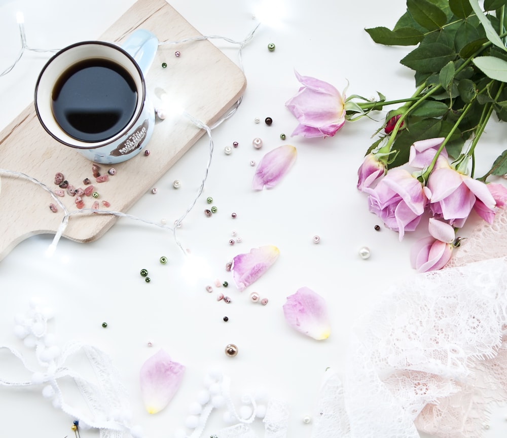 white ceramic mug filled with black liquid on beige chopping board beside pink roses on white surface