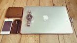 A collection of personal items neatly arranged on a wooden surface. Items include a closed silver laptop with a recognizable logo, a silver wristwatch resting on the laptop, a brown leather wallet, a white smartphone, a black portable charger, and a pair of wired earphones.