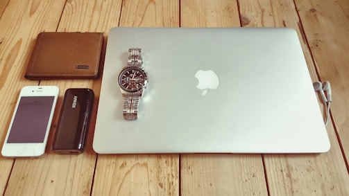 A collection of personal items neatly arranged on a wooden surface. Items include a closed silver laptop with a recognizable logo, a silver wristwatch resting on the laptop, a brown leather wallet, a white smartphone, a black portable charger, and a pair of wired earphones.