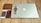 A collection of personal items neatly arranged on a wooden surface. Items include a closed silver laptop with a recognizable logo, a silver wristwatch resting on the laptop, a brown leather wallet, a white smartphone, a black portable charger, and a pair of wired earphones.