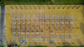 Aerial view of an industrial electrical substation with a complex grid of wires and equipment arranged in neat rows. The ground surface is a sandy beige, contrasting with the silver and dark metal structures. Lush greenery borders the facility at the top edge.