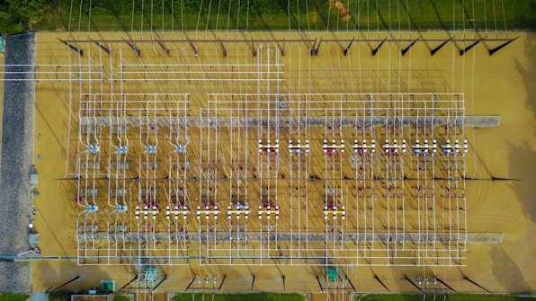 Aerial view of an industrial electrical substation with a complex grid of wires and equipment arranged in neat rows. The ground surface is a sandy beige, contrasting with the silver and dark metal structures. Lush greenery borders the facility at the top edge.