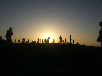 Sunset view from a hilltop with silhouettes of people dancing joyfully around a campfire.