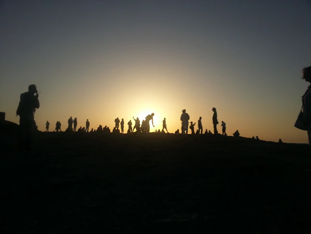 Sunset view from a hilltop with silhouettes of people dancing joyfully around a campfire.