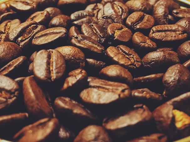 A close-up view of roasted coffee beans with a rich, dark texture. The beans have a glossy finish and varying shades of brown, ranging from dark chocolate to a lighter, earthy hue.