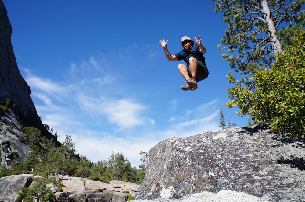man jumping of rock formation near green tree at daytime