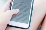 A person is holding a smartphone that displays the text of the Book of Genesis. The background appears soft and out of focus, suggesting a relaxed setting.