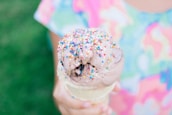 A multicolored sprinkle-covered chocolate ice cream scoop held in a cone. The background features a blur of vibrant colors, likely a child's shirt. The ice cream appears partly melted, suggesting a warm day.