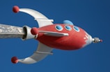 A stylized, retro-futuristic rocket with a red body and silver accents is depicted against a clear blue sky. The rocket features round blue windows and red spheres on its silver fins.