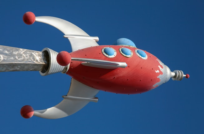 A stylized, retro-futuristic rocket with a red body and silver accents is depicted against a clear blue sky. The rocket features round blue windows and red spheres on its silver fins.