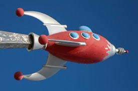 A stylized, retro-futuristic rocket with a red body and silver accents is depicted against a clear blue sky. The rocket features round blue windows and red spheres on its silver fins.