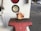 A cartoonish figurine dressed in a biker outfit stands on a red metal surface, likely part of a piece of machinery or equipment. The background includes parts of a utility vehicle, such as a lock and circular lights in black and red casings. The scene is outdoors with a parking lot visible in the distance.